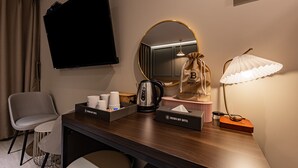 A hotel room with a flat-screen TV, a round mirror, a desk with a coffee maker, a tissue box, a lamp, and a chair. - Brown Dot Hotel (Jecheon)