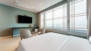 A modern hotel room with a large bed, a flat-screen TV mounted on the wall, a desk with a chair, and a large window offering a view of the city. - Brown Dot Hotel (Jecheon)