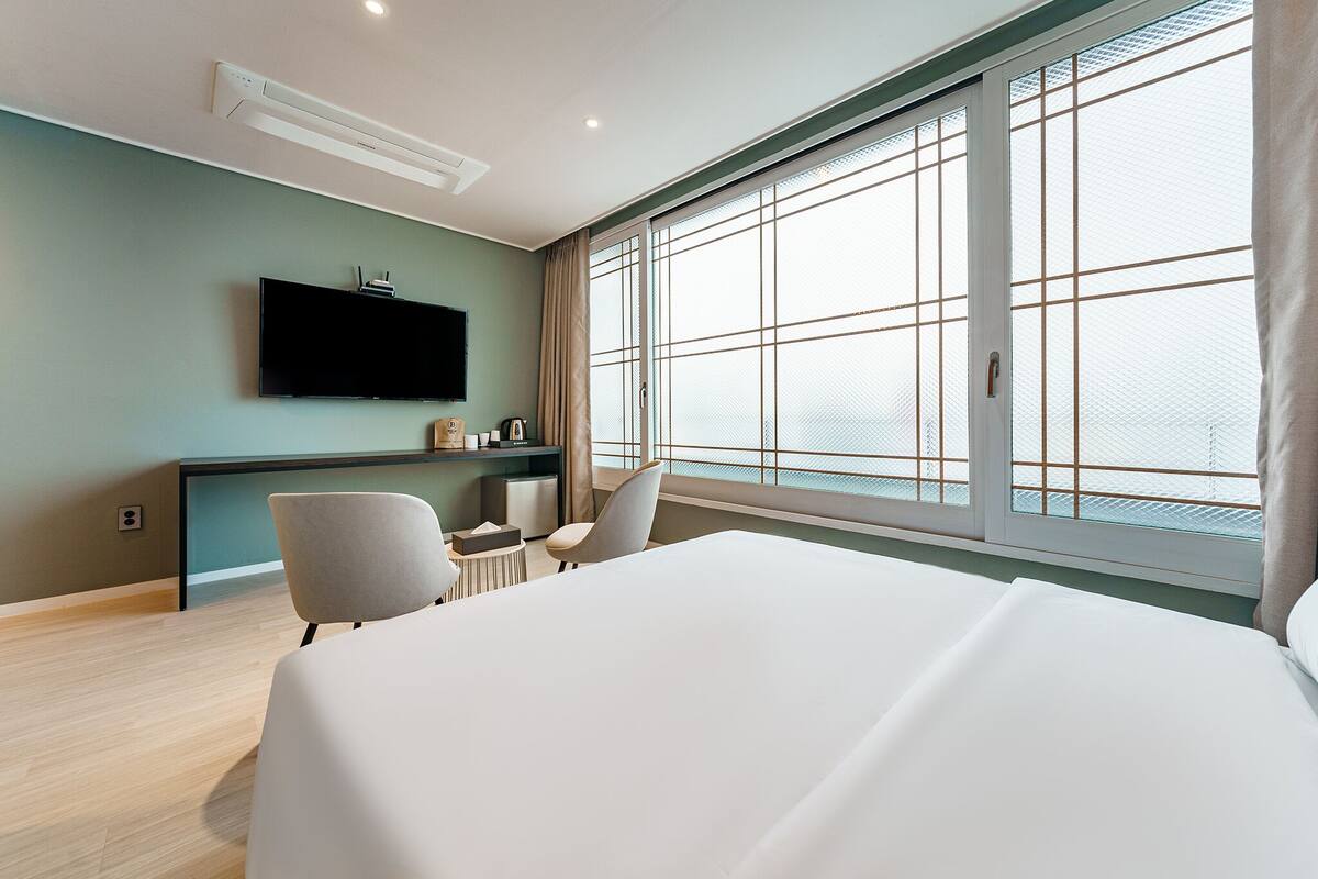 A modern hotel room with a large bed, a flat-screen TV mounted on the wall, a desk with a chair, and a large window offering a view of the city.