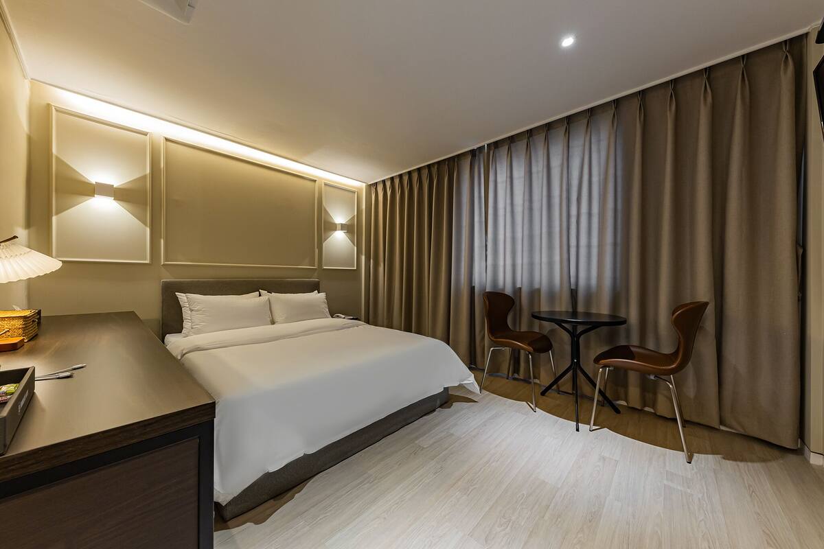 A hotel room with a large bed, a desk with a lamp, two chairs, and a large window with curtains.