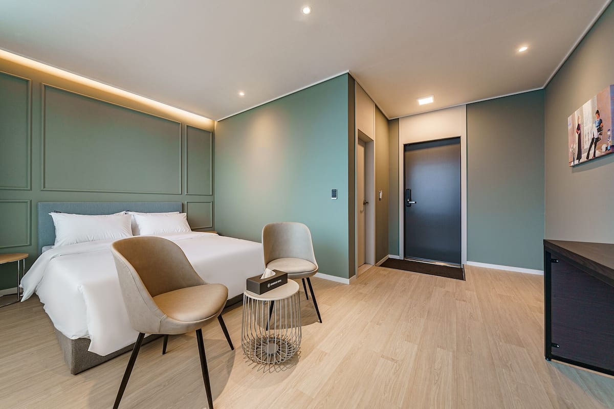 A modern hotel room with a bed, two chairs, a small table, and a door.