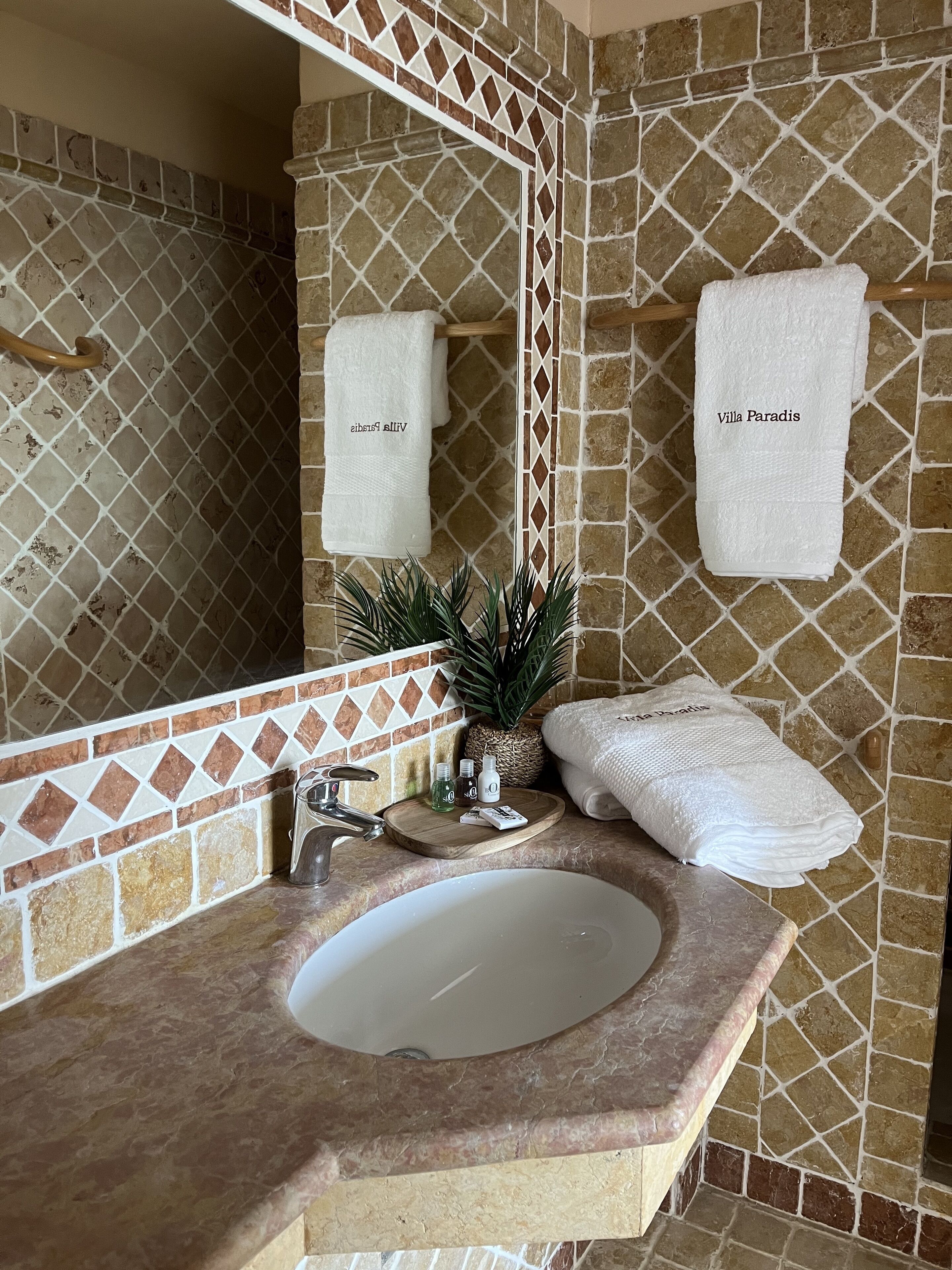Een badkamer met een backsplash van stenen mozaïek, een wastafel, een spiegel en twee handdoeken met de tekst 'Villa Paradis'.