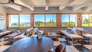 A spacious room with large windows offering a view of the sea and greenery. There are multiple seating areas with wooden furniture and a central table. - Villa Paradis (Roquebrune-sur-Argens)