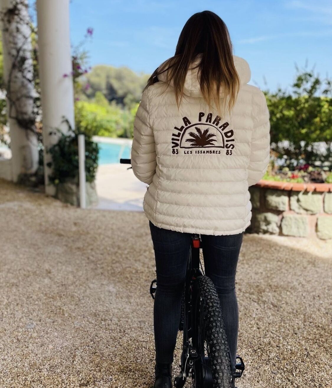 Una persona montando en bicicleta al aire libre, vistiendo una chaqueta blanca con un logotipo y texto en la parte trasera.