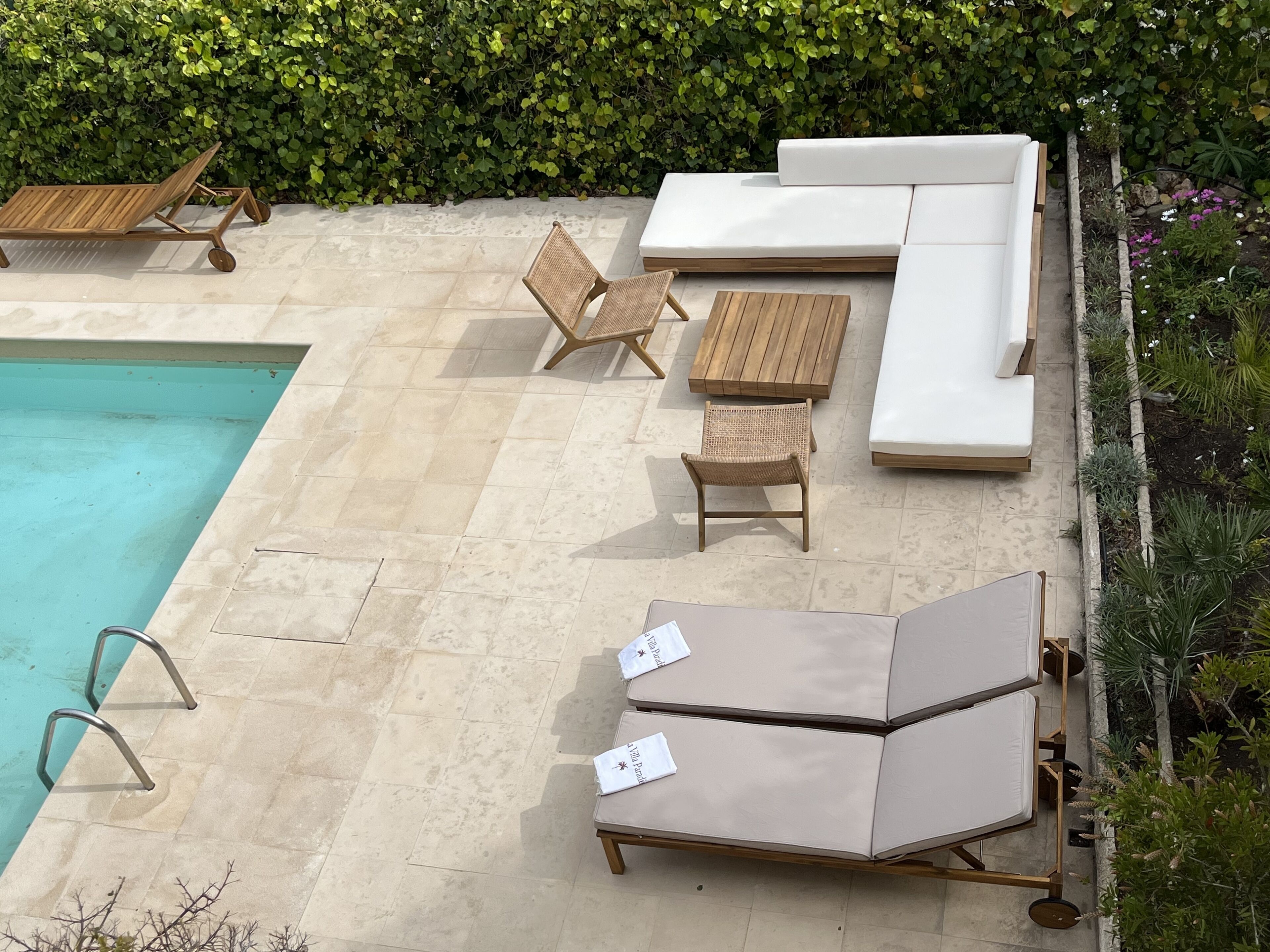 Zona junto a la piscina con una tumbona, una mesa de madera, una silla de mimbre y un sofá con cojines.