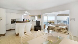 Une cuisine moderne avec des meubles blancs, un coin repas doté d’une table en verre et une vue sur la mer.