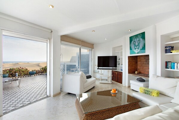 A modern living room with a large TV, white sofas, a glass coffee table, and a view of the desert.