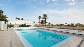 Une piscine rectangulaire, bordée d’une terrasse carrelée, entourée de palmiers et avec un bâtiment blanc en arrière-plan.