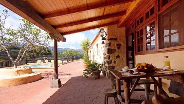 Eine Holzpergola mit einer Steinmauer, einem Pool und einem Tisch mit Obst und Getränken.