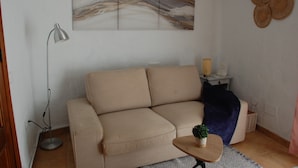 A living room with a beige sofa, a floor lamp, a small table with a plant, a wall art piece, and a decorative wall hanging.