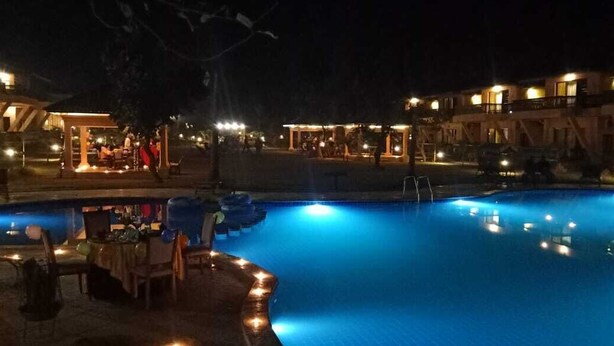 A nighttime swimming pool area with lounge chairs and a bar, surrounded by illuminated buildings. - The Pench International (Kurai)