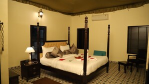 A four-poster bed with a headboard, a bedside table with a lamp, a chair, and a small table. - The Pench International (Kurai)