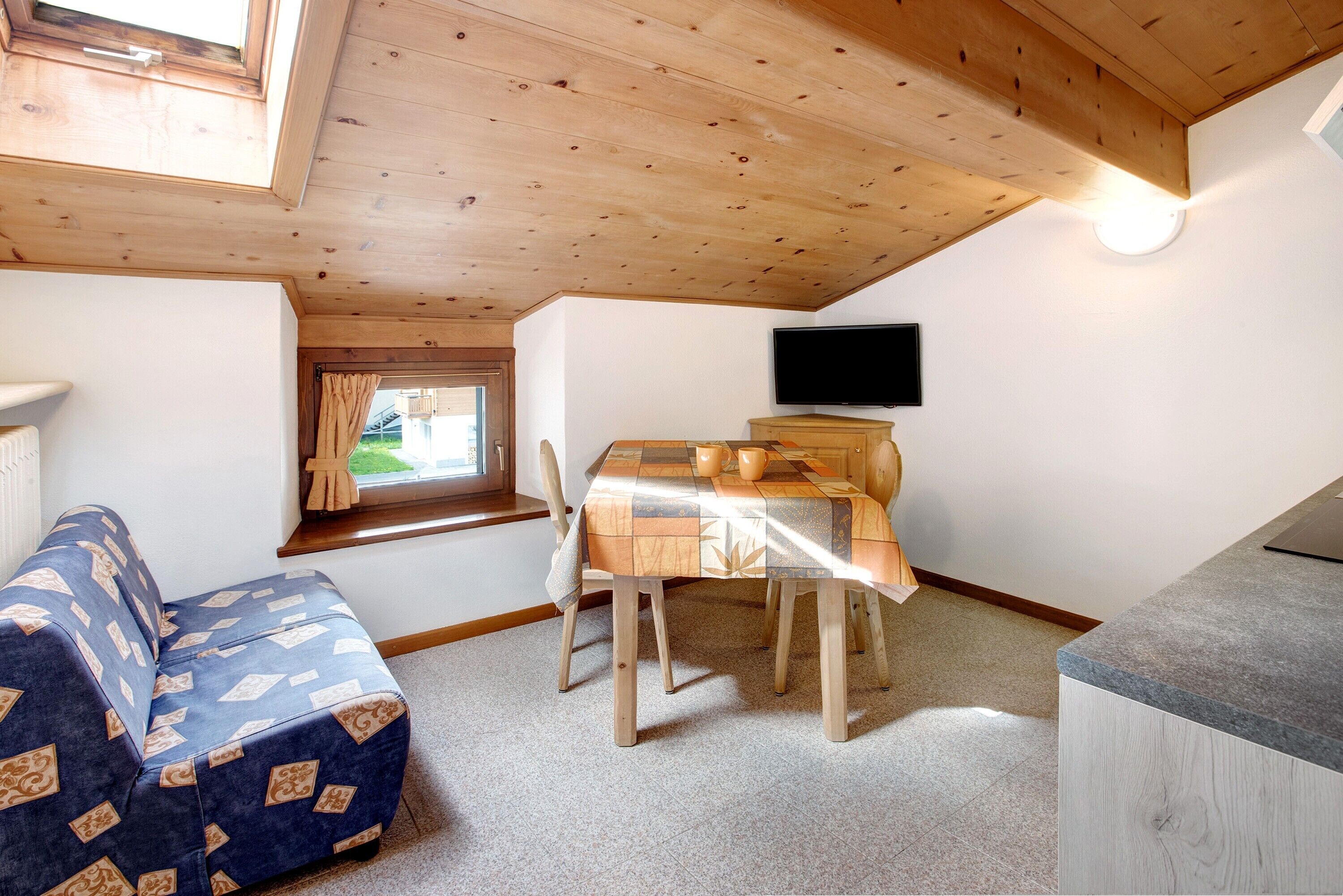 Ein gemütliches Zimmer mit Holzbalkendecke, einem Esstisch mit Stühlen, einem an der Wand montierten Fernseher und einem Fenster mit Vorhängen.