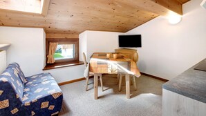 A cozy room with a wooden ceiling, a dining table with chairs, a television mounted on the wall, and a window with curtains.