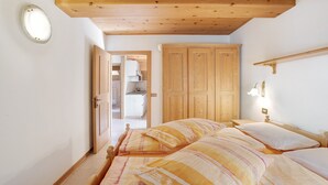A bedroom with a wooden ceiling, a bed with striped bedding, a wardrobe, and a small kitchen visible through an open door.