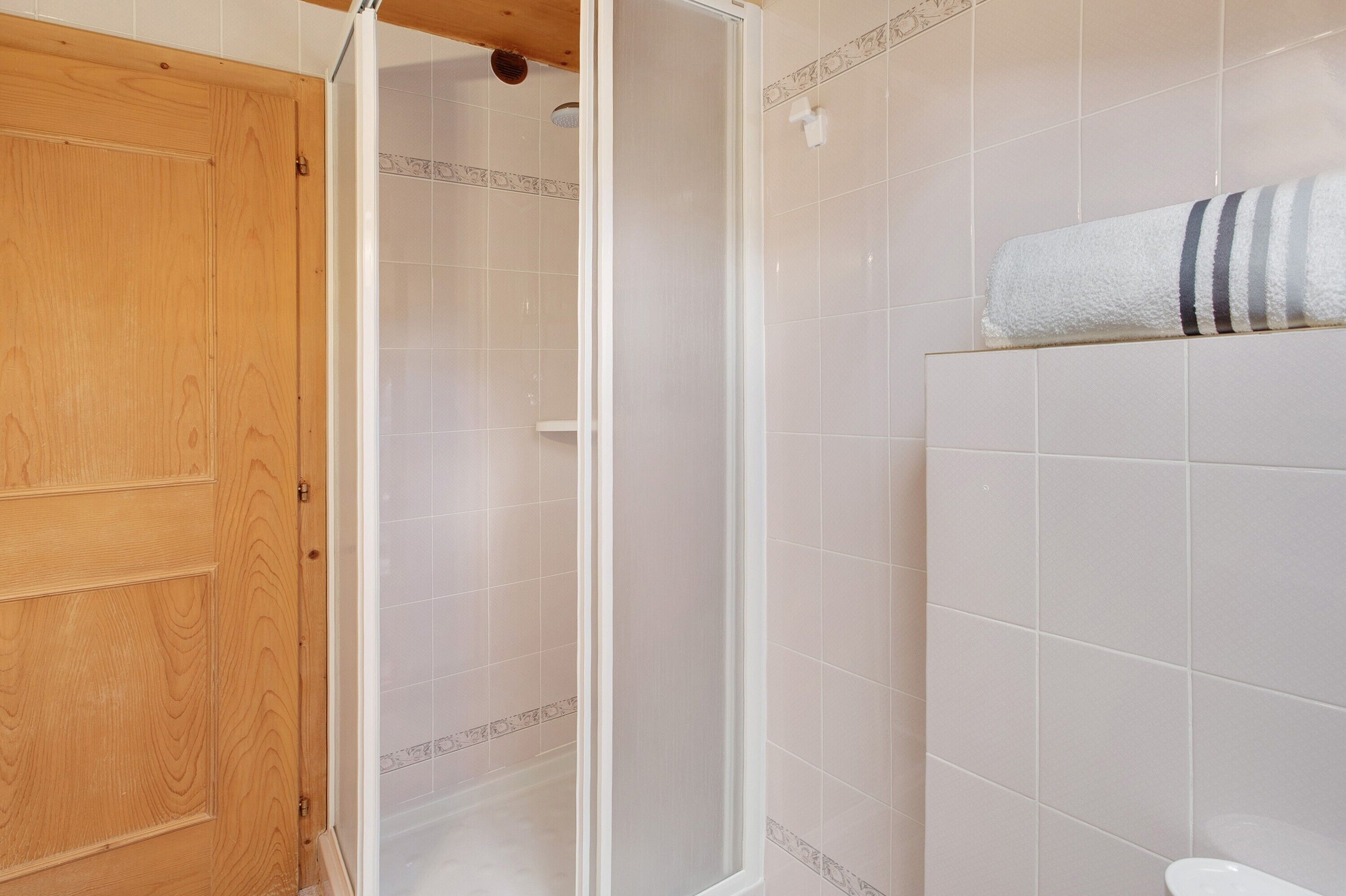 A bathroom with a wooden door, a shower enclosure, and a towel on a shelf.