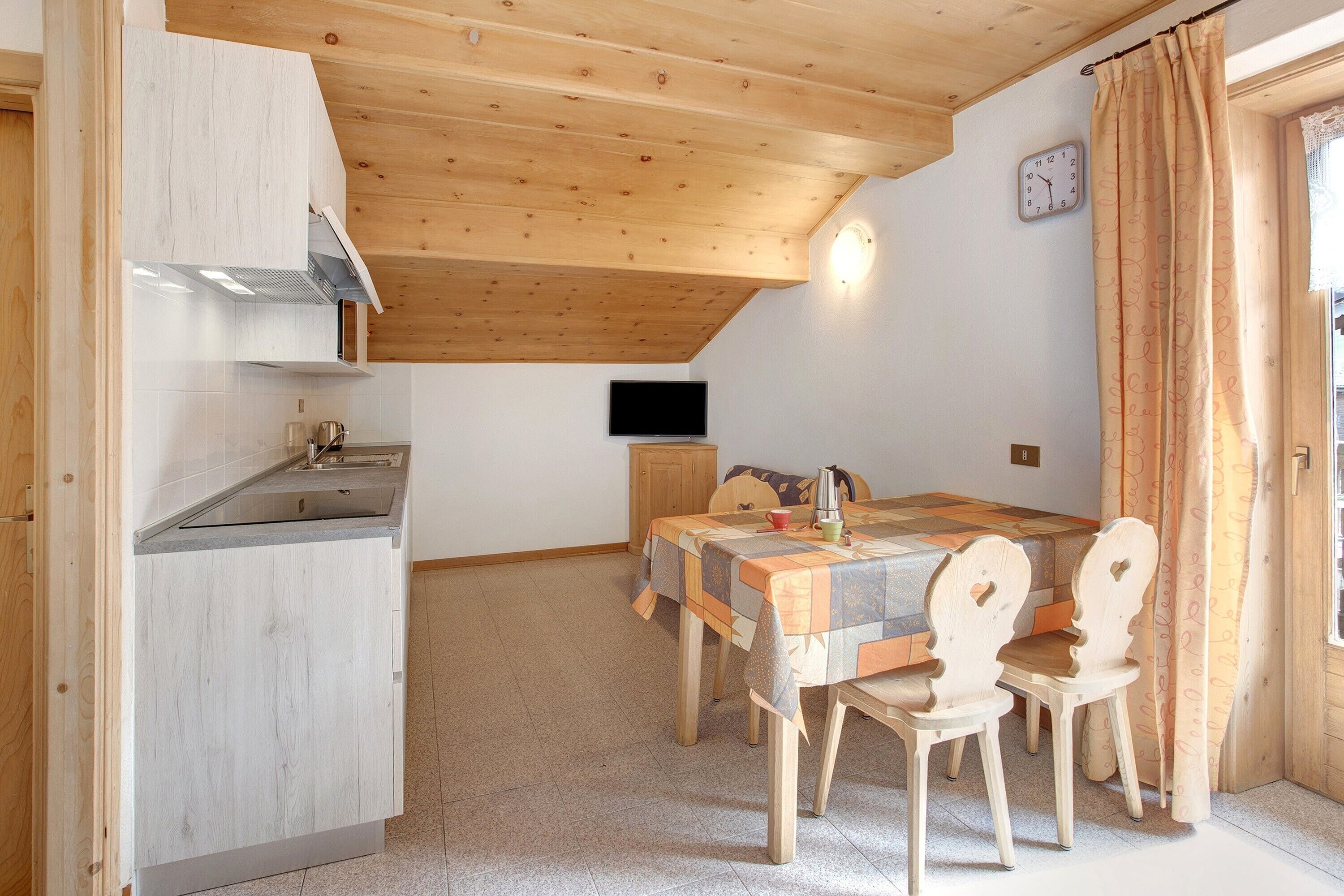 Una cocina con armarios de madera, una mesa de comedor con sillas, un televisor y un reloj en la pared.