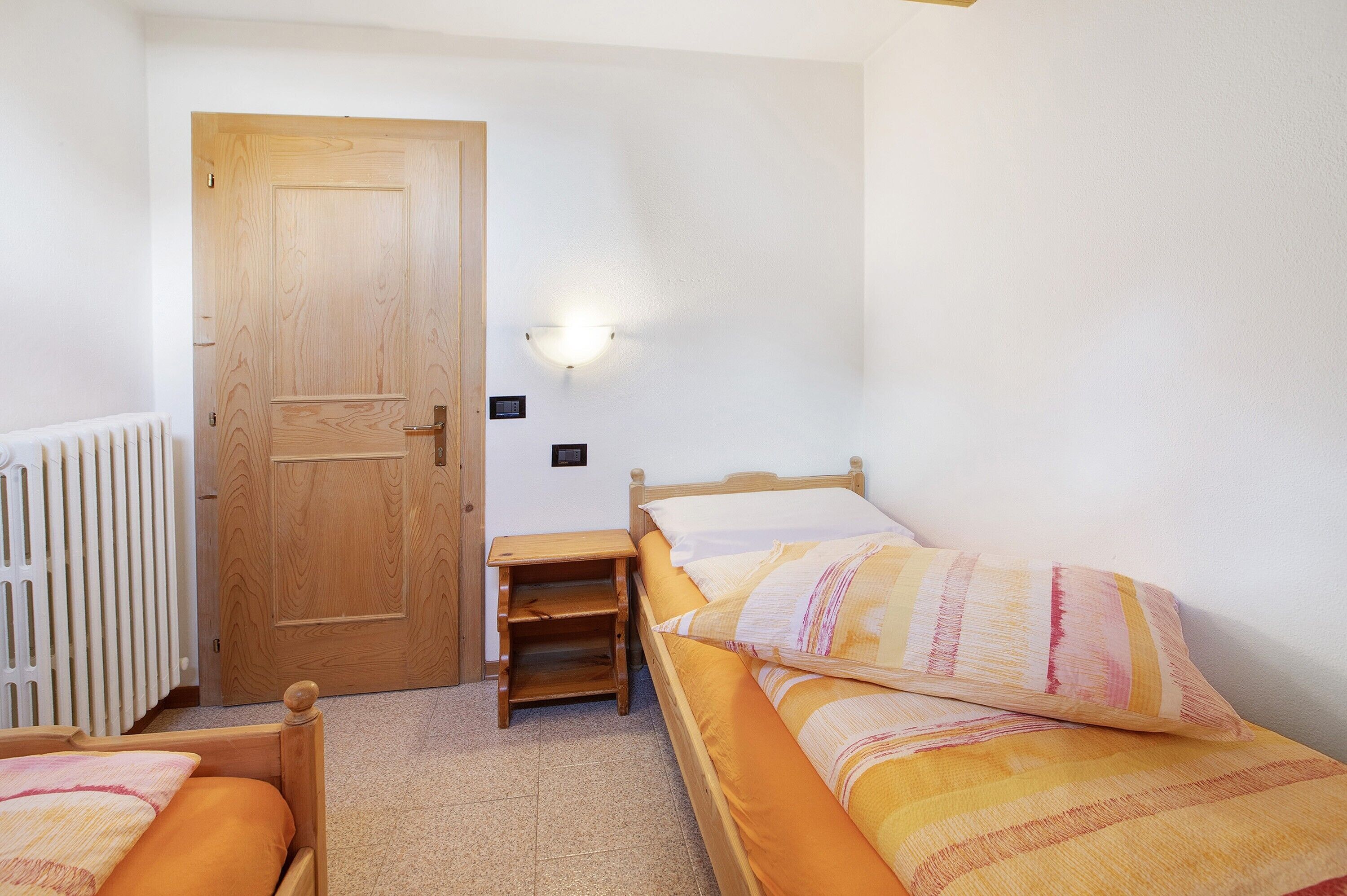 Une chambre avec deux lits simples, une porte en bois, une table de chevet, une lampe fixée au mur et un radiateur.