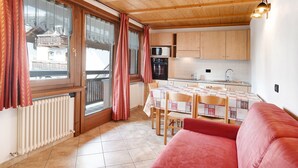 Ein gemütliches Zimmer mit Holzboden, einem Esstisch mit Stühlen, einem roten Sofa, einer Küchenzeile mit Schränken und Geräten sowie Blick durch große, mit Vorhängen drapierten Fenster.