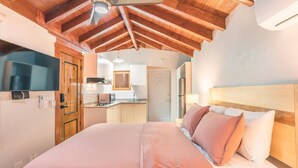 Une chambre avec un plafond en bois, un lit avec une literie rose et blanche, une télévision sur le côté gauche et, au fond, une petite cuisine.