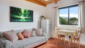 Een kamer met een bank, een tafel met stoelen, een koelkast, een raam met gordijnen, en een schilderij aan de muur.