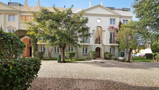 A large building with a cobblestone courtyard, arched doorways, and a balcony with flowers. - Apartment 'Old Village T2 Duplex' with Shared Pool, Wi-Fi and Air Conditioning (Quarteira)