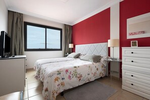 Un dormitorio con cama, cómoda, mesita de noche, lámpara, televisor y ventana con cortinas.