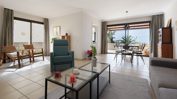 Un salon comprenant un canapé, un fauteuil, une table basse en verre, une table à manger avec des chaises, et une vue dégagée grâce à de grandes fenêtres.