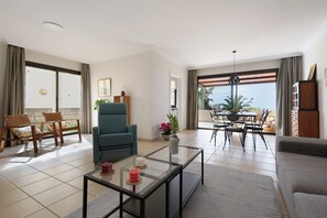 Una sala de estar con sofá, sillón, mesa de centro de vidrio, mesa de comedor con sillas y vistas amplias a través de grandes ventanales.