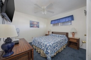 Une chambre à coucher avec un lit, un ventilateur de plafond, un téléviseur fixé au mur, une fenêtre avec des rideaux bleus et un tableau accroché au mur.