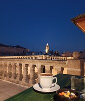 Un balcón con una taza de café y un bol de fruta, con vistas a una torre y un paisaje urbano al anochecer.