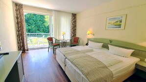 Ein Hotelzimmer mit Bett, Schreibtisch, Stuhl, Tisch mit Lampe und Blick ins Grüne durch das Fenster.