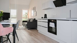 A modern kitchen with white cabinetry, a built-in oven, and a black range hood. There is a dining area with a table, chairs, and a vase of flowers. - Apartment 2 Teefabrik - Living at the Norder Tief (Lorup)