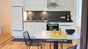 Une cuisine moderne avec un plan de travail blanc, des appareils en acier inoxydable et un crédence noire. On y trouve une table à manger avec deux chaises et un saladier posé dessus.