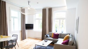 Un salon moderne avec un canapé, une table basse et des fauteuils. Un téléviseur est fixé au mur et il y a une fenêtre avec des rideaux.