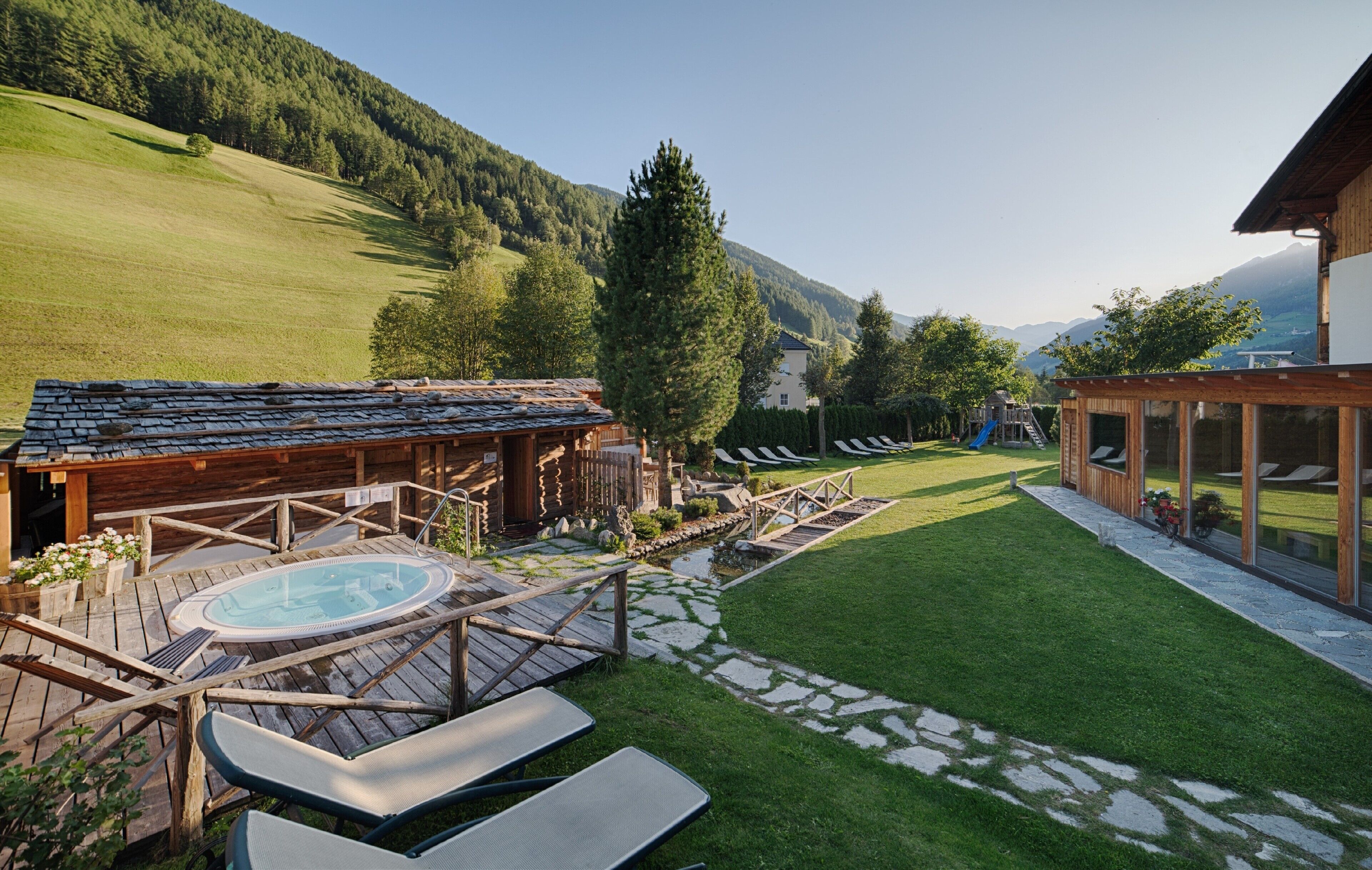 A wooden cabin with a hot tub, a grassy yard, and a mountain backdrop.