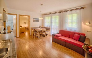 A modern living room with a wooden floor, a red sofa, a dining table with chairs, and a kitchen area with a sink and cabinets.