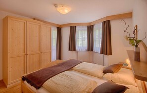 A bedroom with a large wardrobe, a bed with two pillows, a bedside table with a lamp, and a window with curtains.