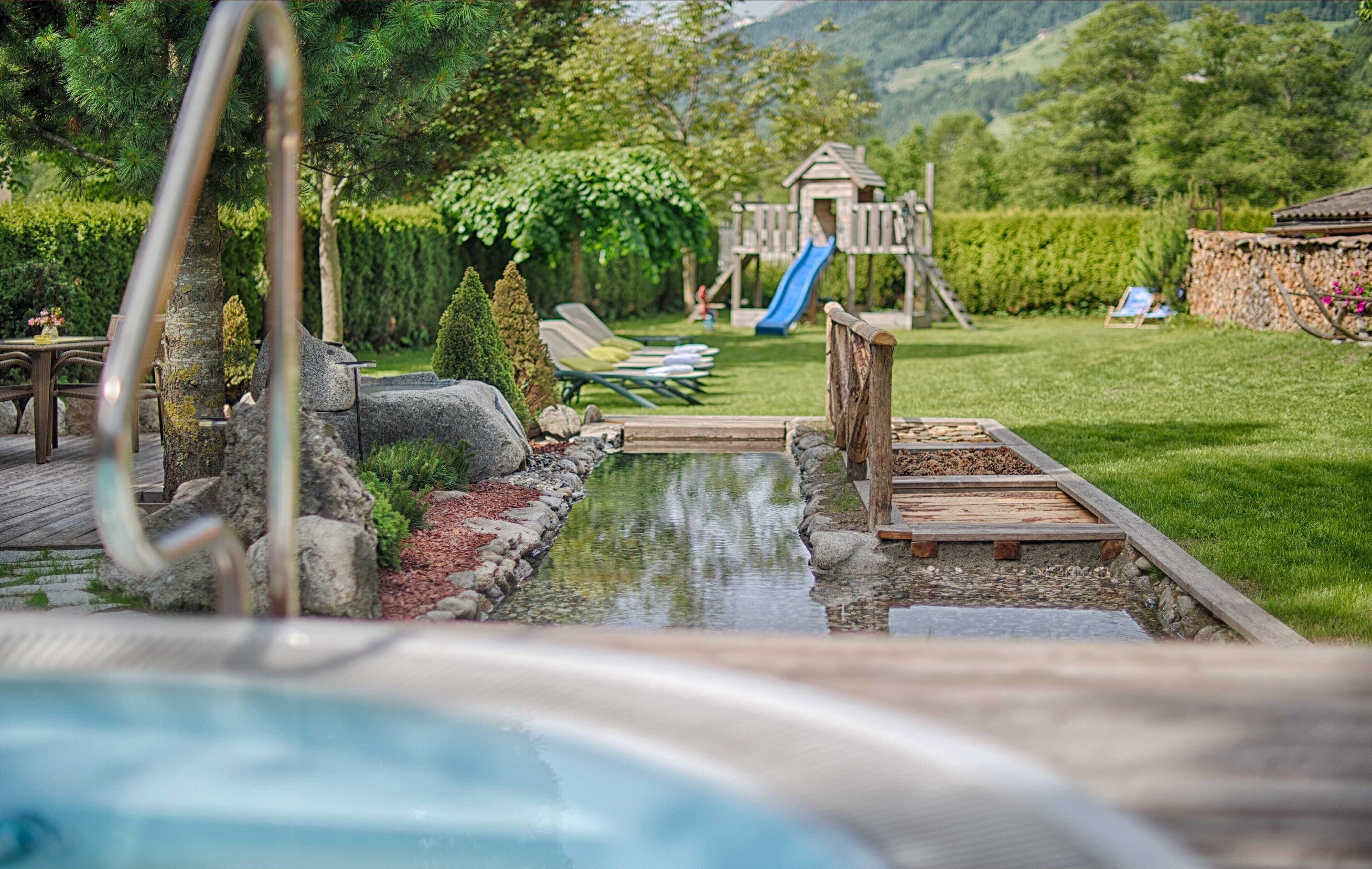 Ein Pool mit Holzterrasse, Liegestühlen, einer Rutsche und einer Spielanlage in einem angelegten Garten.