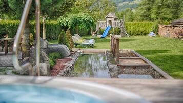 Ein Schwimmbad mit Holzterrasse, Liegestühlen, einer Rutsche und einem Spielplatz im Hintergrund.