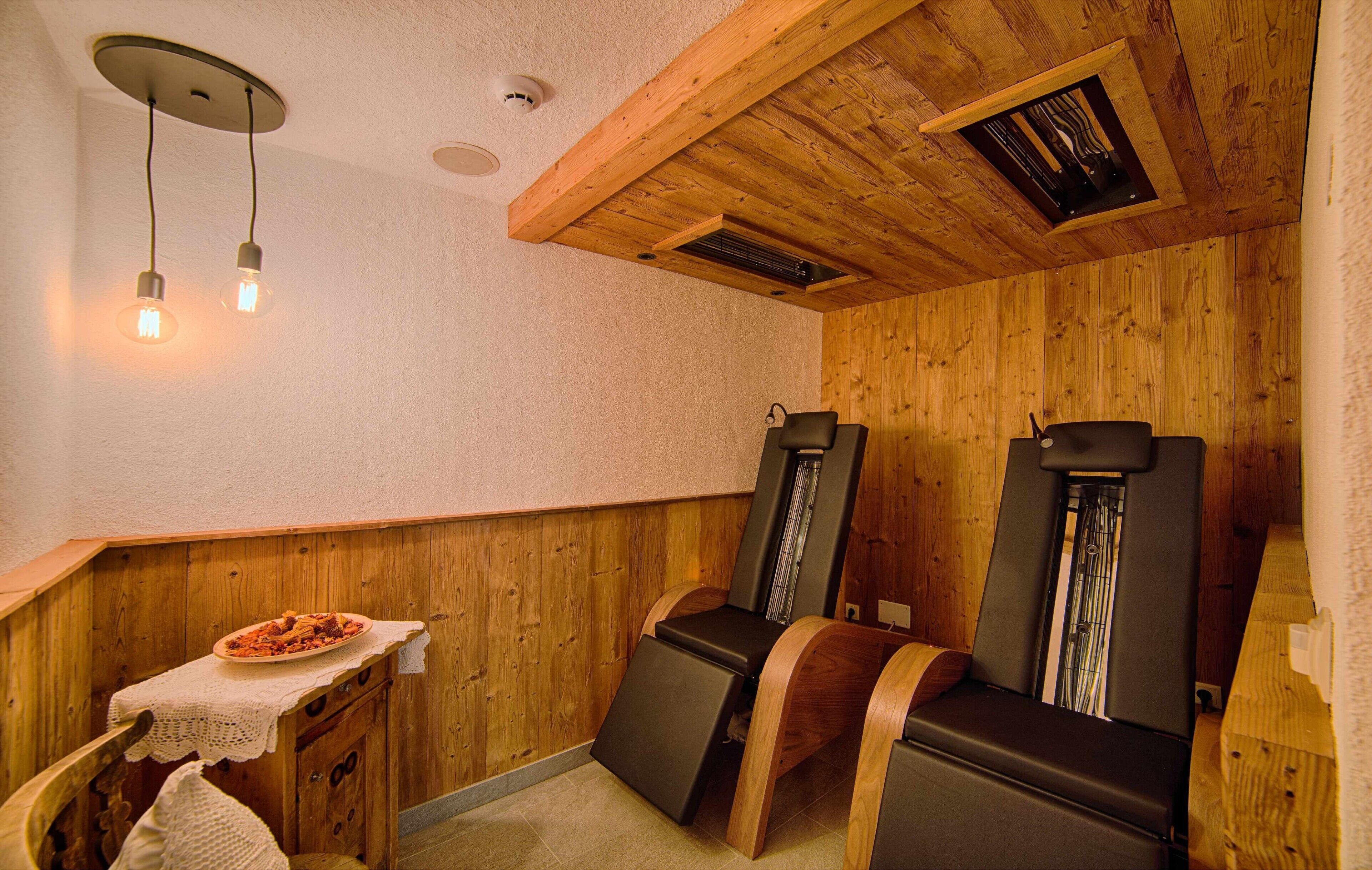 A sauna room with wooden walls, two massage chairs, a small table with a towel, and a ceiling vent.