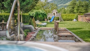 Ein Schwimmbad mit Holzterrasse, Liegestühlen, einer Rutsche und einem Spielplatz im Hintergrund.