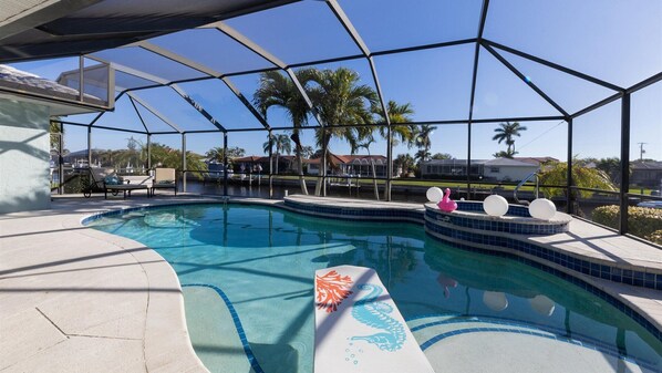 A swimming pool with a glass enclosure, lounge chairs, and a view of palm trees and houses in the background.