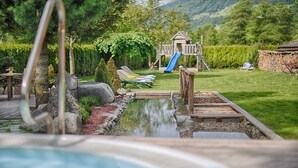 A swimming pool with a wooden deck, lounge chairs, a slide, and a playground in the background.