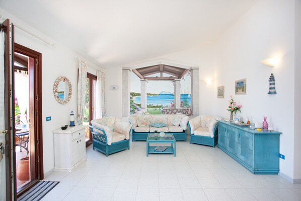 A living room with a view of the sea, a blue sofa set, a glass coffee table, and a white cabinet.