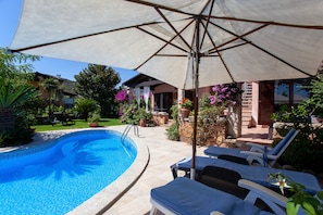 A swimming pool with lounge chairs and an umbrella, surrounded by potted plants and a stone wall.