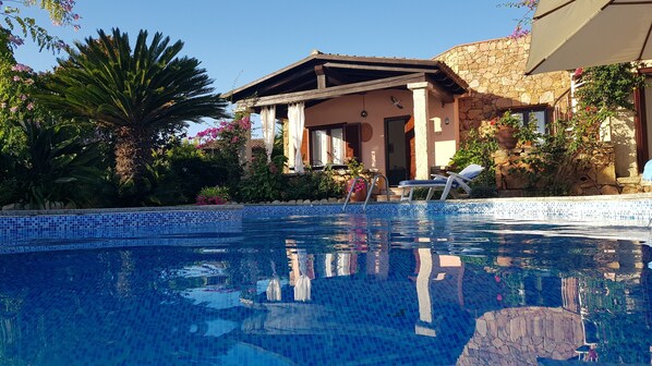 Ein Swimmingpool mit einer Fliesenverkleidung, eine Liege und ein Steinhaus mit einem Holzdach.