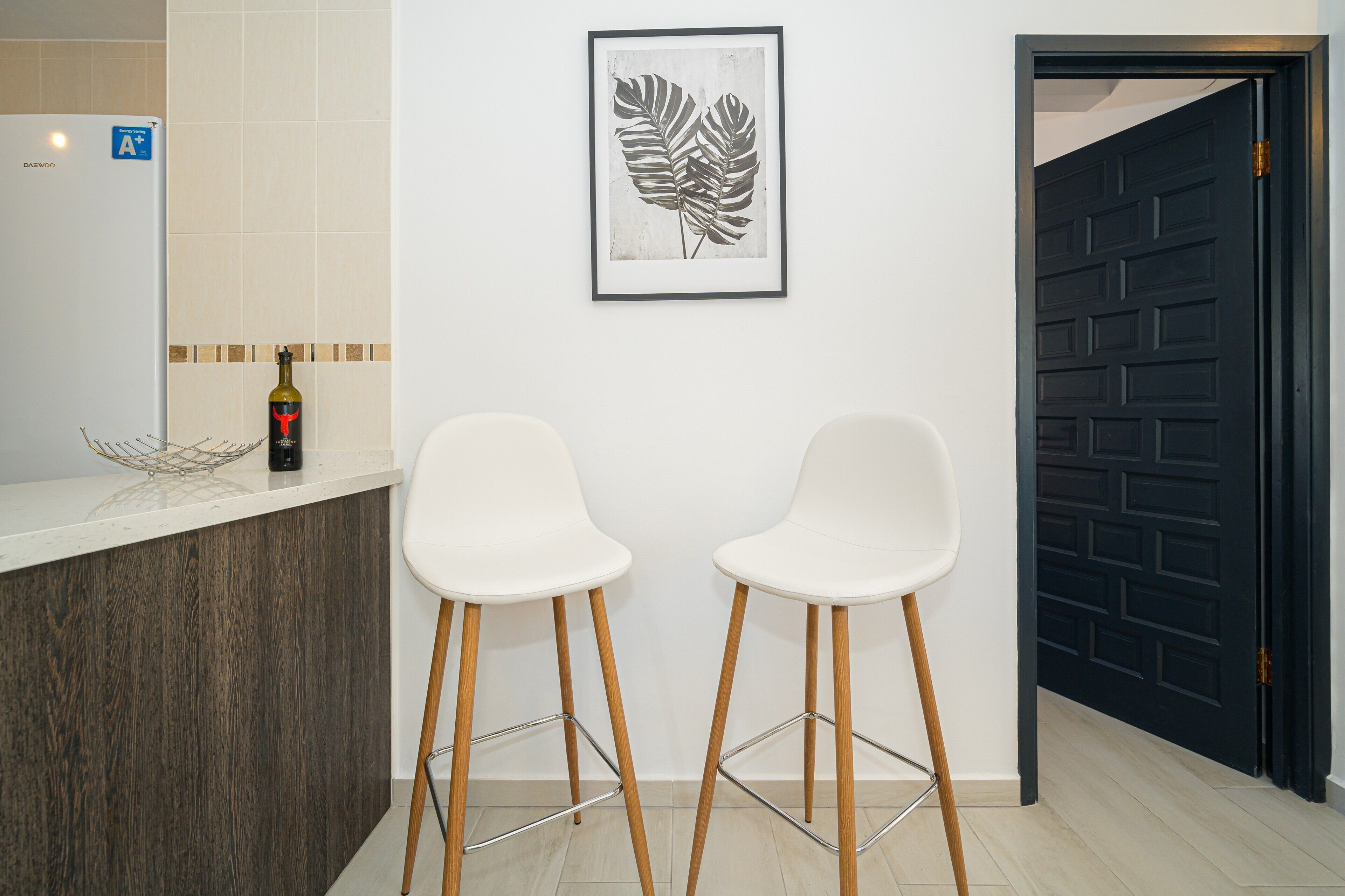 Una cocina con encimera blanca, dos taburetes de bar blancos, una obra de arte con hojas enmarco y una puerta negra.