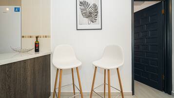 Una cocina con encimera blanca, dos taburetes de bar blancos, una obra de arte con hojas enmarco y una puerta negra.