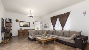 Una sala de estar con un sofá seccional, una mesa de centro de madera, una araña y una zona de cocina con armarios y un refrigerador.